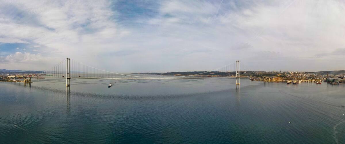A large suspension bridge spanning a calm body of water with a cityscape in the background.