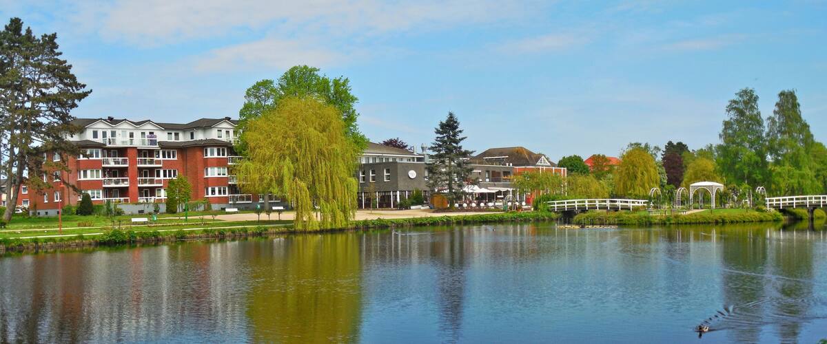A calm lake with a wooden bridge, a gazebo, and buildings in the background.