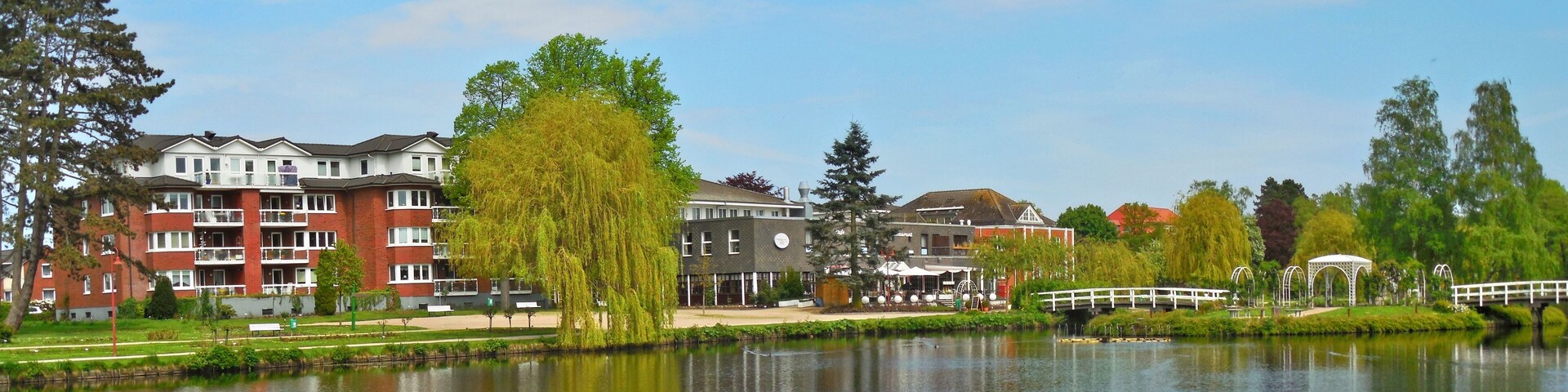 A calm lake with a wooden bridge, a gazebo, and buildings in the background.