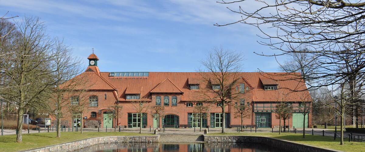 A red-brick building with a clock tower, a pond, and leafless trees.