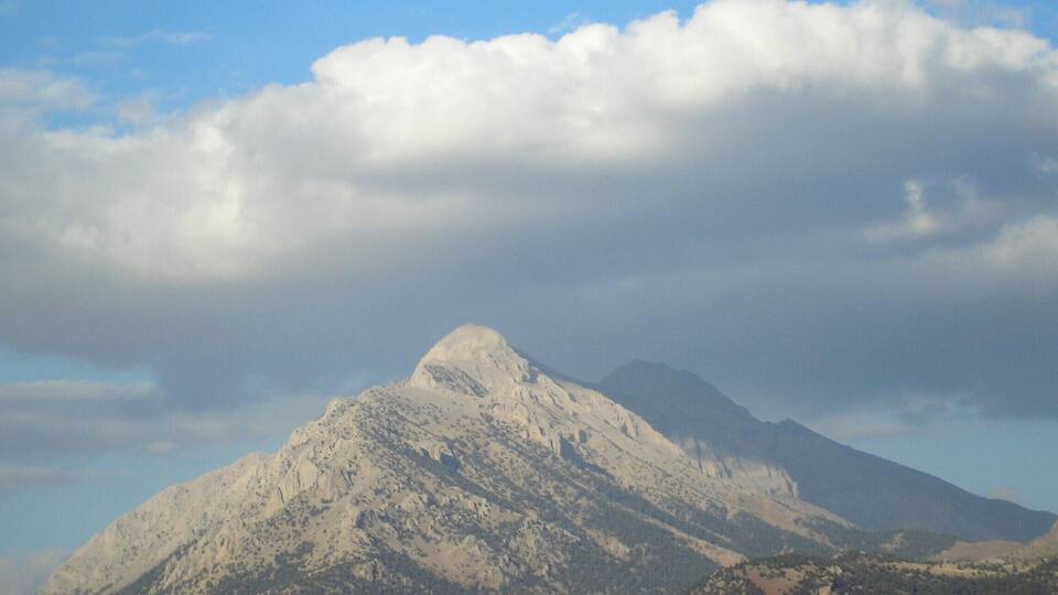 A snow-capped mountain with a forested foreground and a partly cloudy sky.