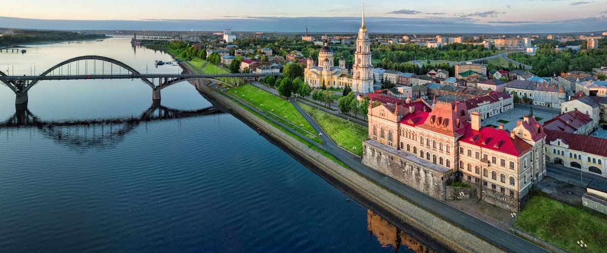 A river with a bridge, a church with a tall spire, and historic buildings with red roofs.