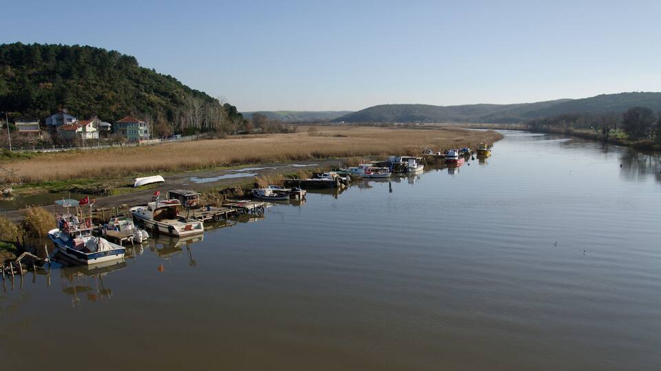 A calm river with boats docked along the shore, surrounded by hills and houses.
