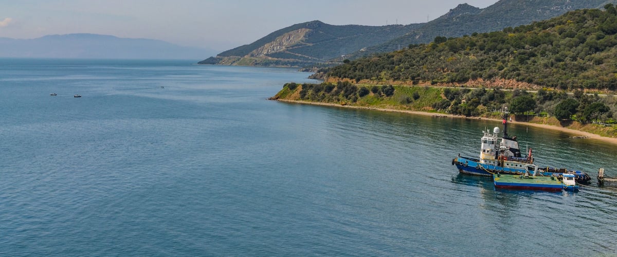 A calm sea with a large boat docked near the shore, surrounded by hills and sparse vegetation.