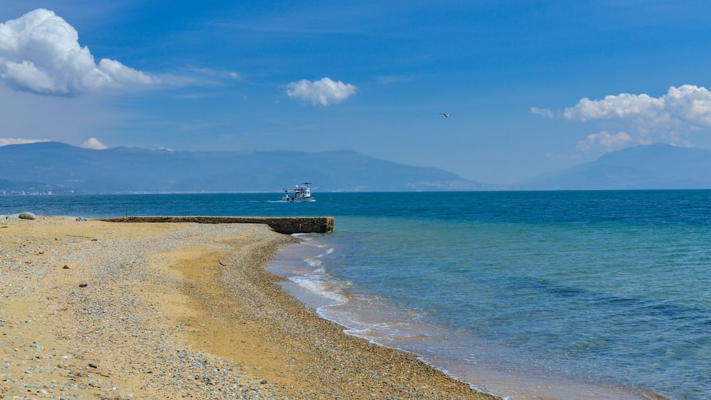 Une plage de galets avec un bateau sur lâeau, des montagnes Ă lâhorizon et un ciel dĂ©gagĂ©.