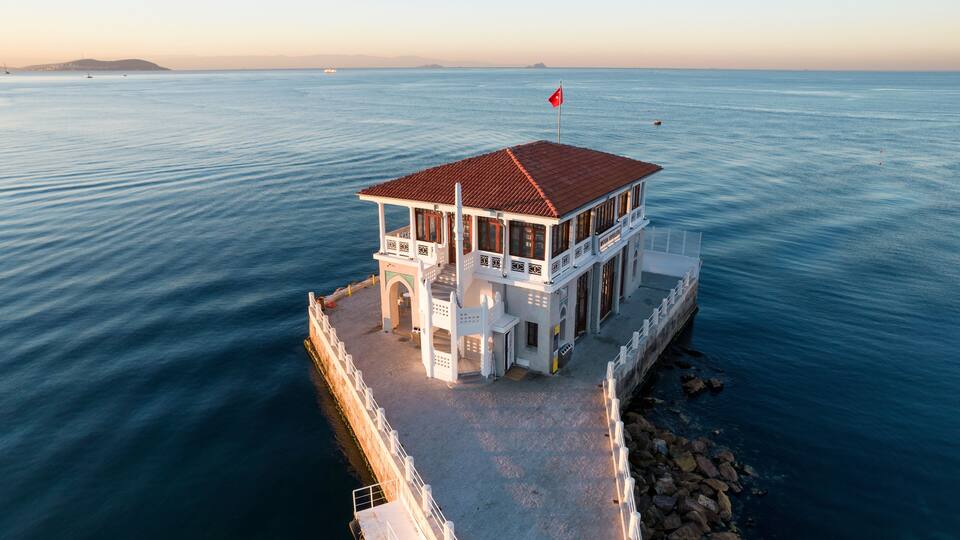 A coastal building with a red roof, white walls, and a flag on top, situated on a rocky shoreline with calm sea in the background.