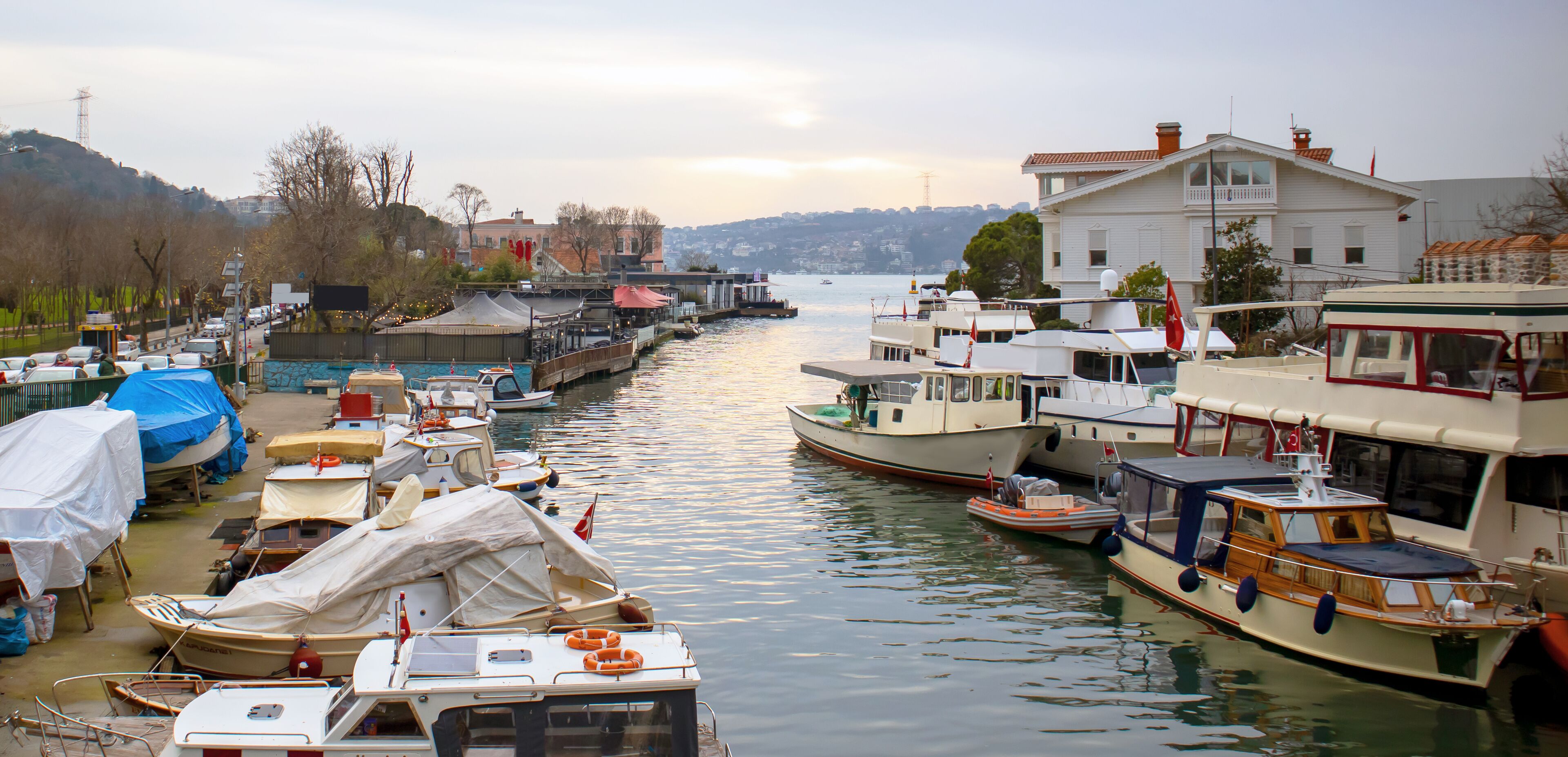 A marina with various boats docked along a river, surrounded by buildings and hills in the background.