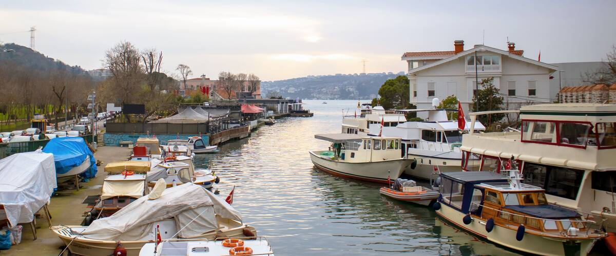 A marina with various boats docked along a river, surrounded by buildings and hills in the background.