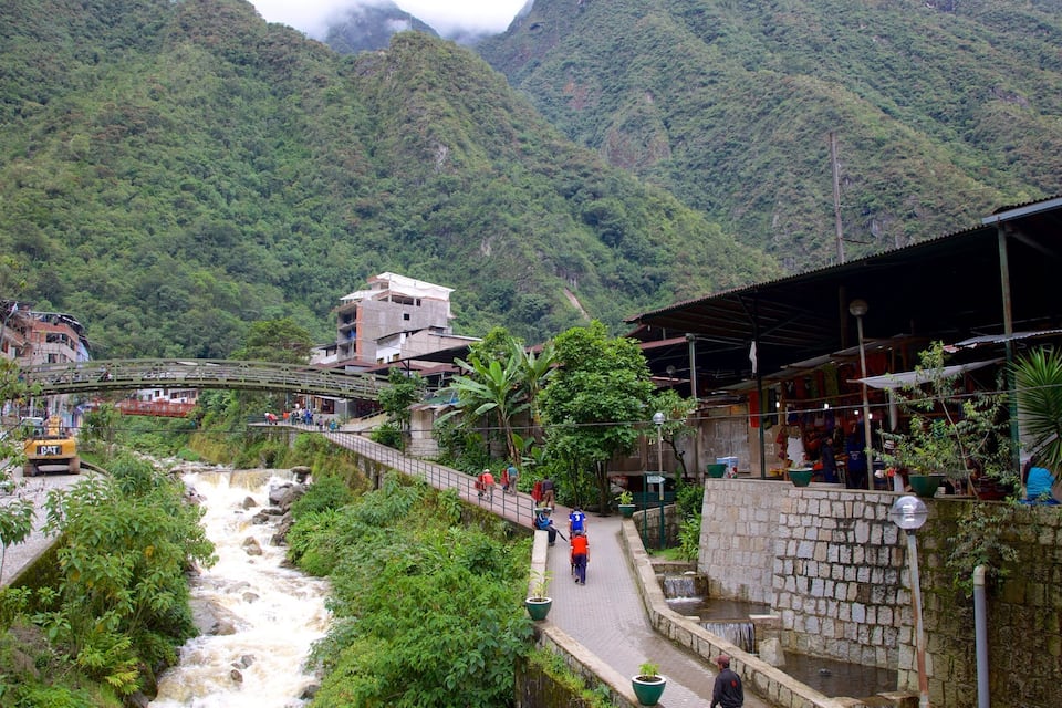 Um rio serpenteando por uma região montanhosa, com uma ponte, edifícios e um calçadão pavimentado.