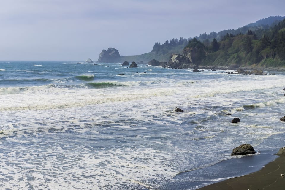 A coastal landscape with waves crashing onto a sandy beach, rocky outcrops, and a forested hillside in the background.