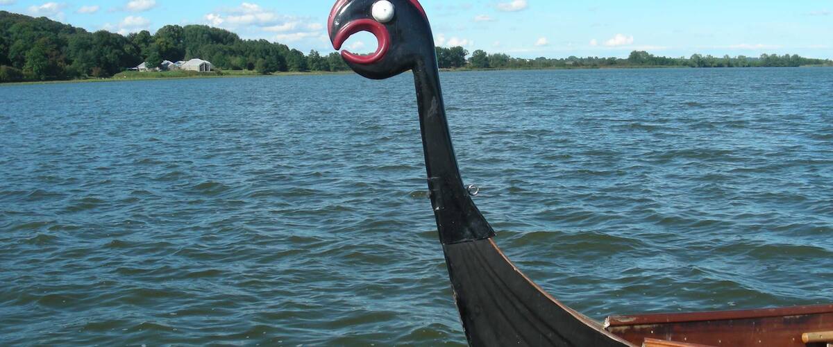 A Viking longship with a curved prow and a red and black head ornament on a calm lake with a forested shoreline.