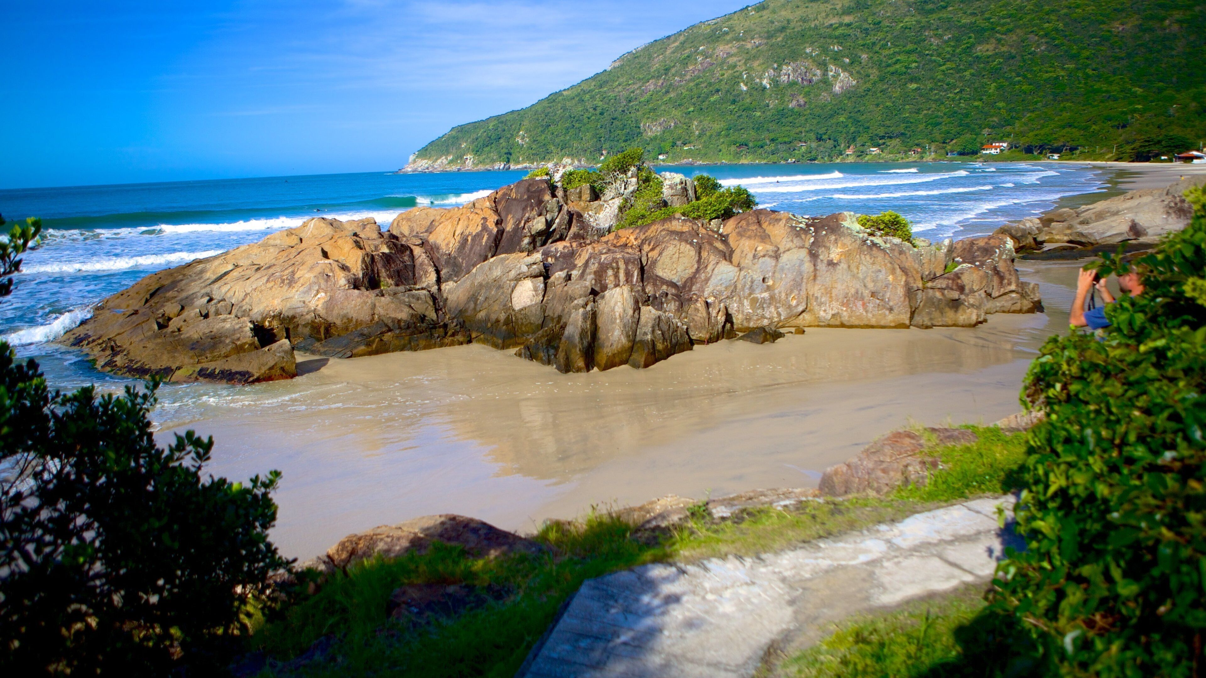 A rocky coastline with waves crashing against the shore, a clear blue sky, and a green hill in the background.