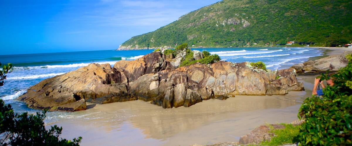 A rocky coastline with waves crashing against the shore, a clear blue sky, and a green hill in the background.