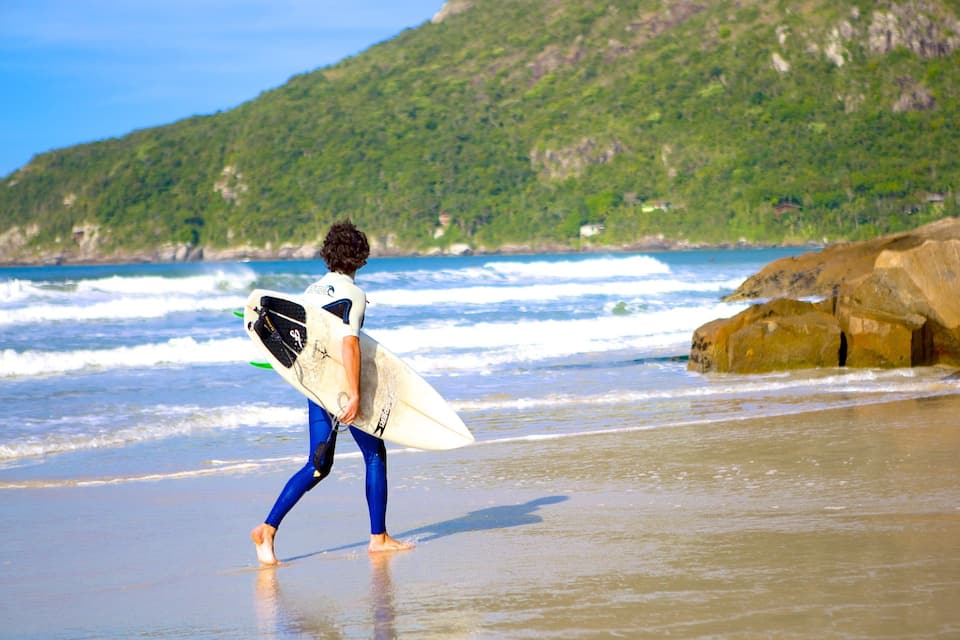A surfer walking on a beach with waves and a mountain in the background.