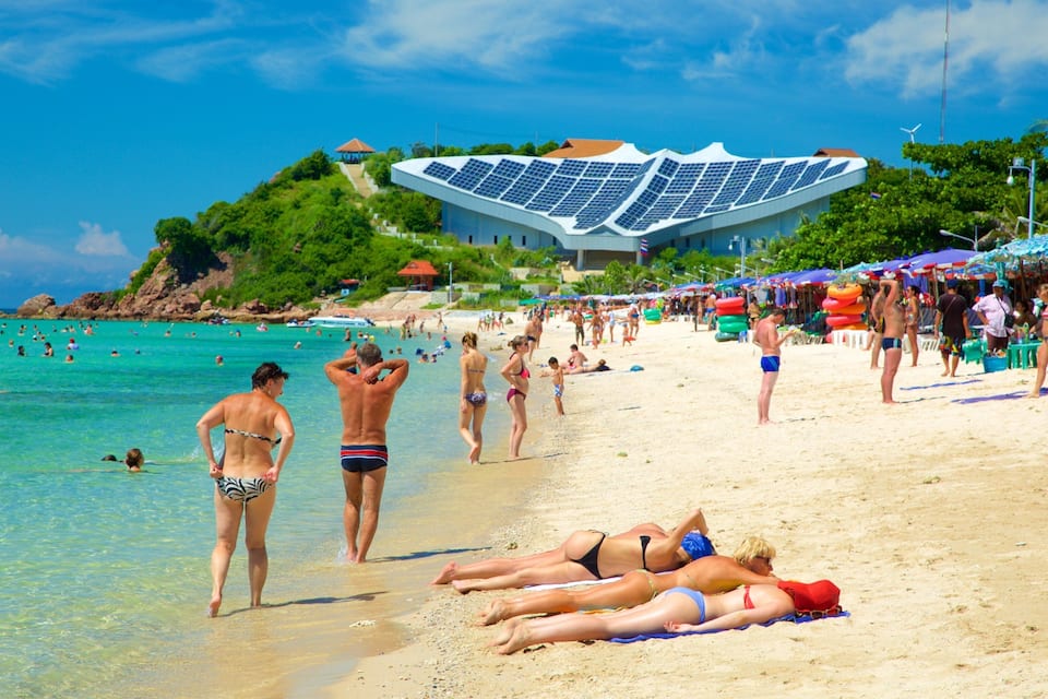 A beach with people sunbathing, swimming, and walking. There is a large building with solar panels in the background.