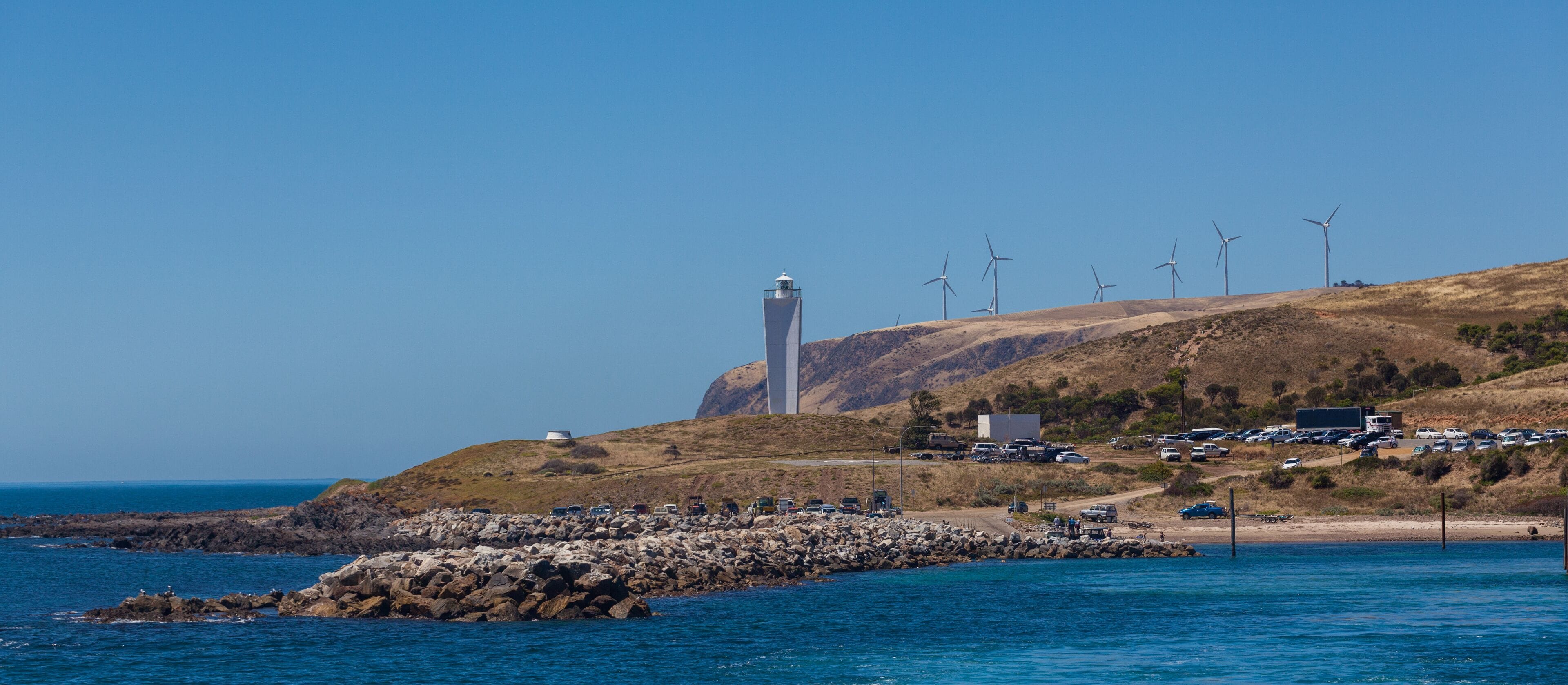 Un paisaje costero con un faro, aerogeneradores y un espigón rocoso.