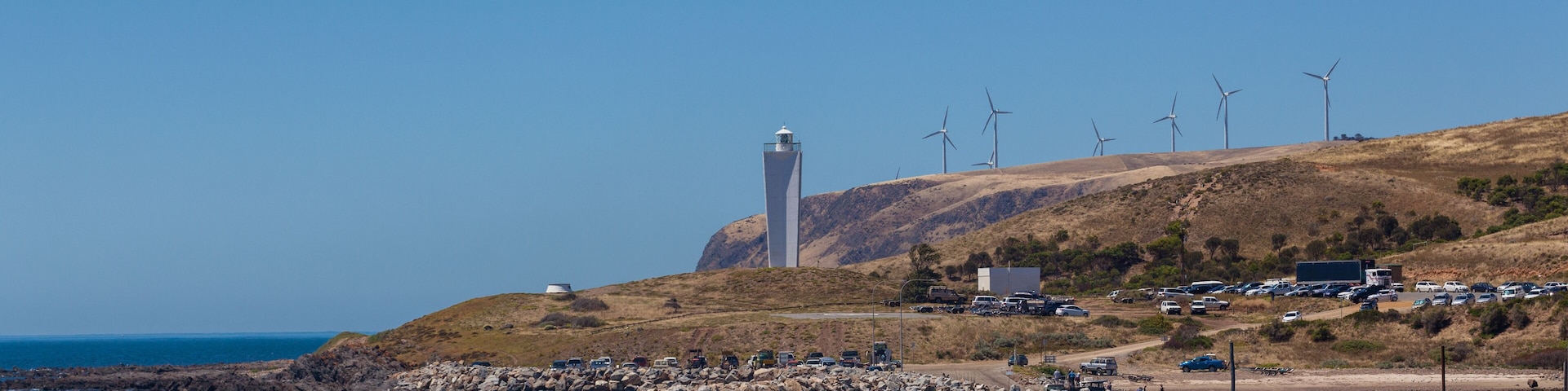 Un paisaje costero con un faro, aerogeneradores y un espigón rocoso.