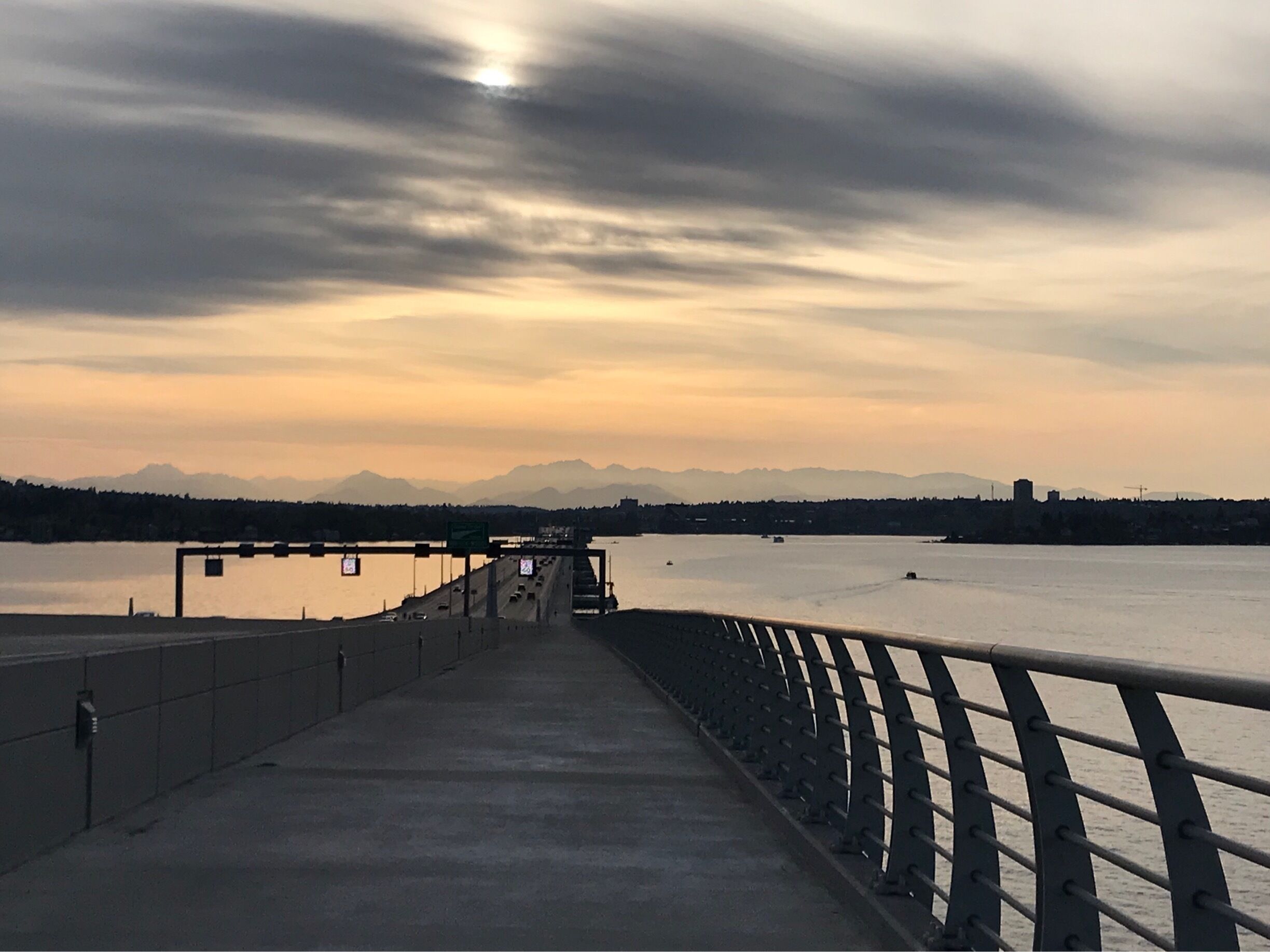 A bridge with a metal railing overlooking a calm river and distant mountains under a cloudy sky.