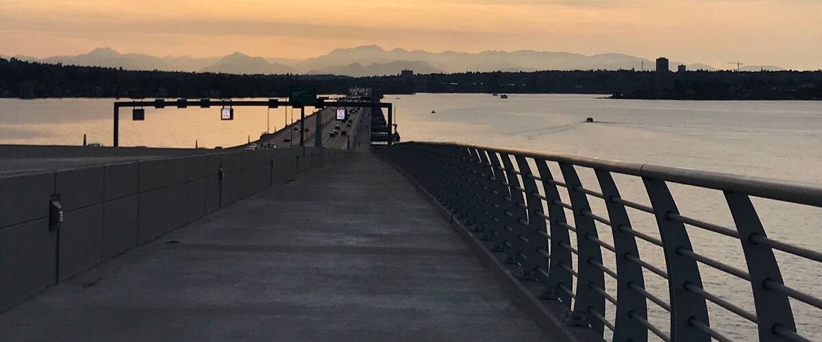 A bridge with a metal railing overlooking a calm river and distant mountains under a cloudy sky.