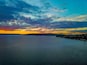 Kystlandskab med solnedgang over vandet, en fjerntliggende kystlinje med bygninger og en himmel med skyer.