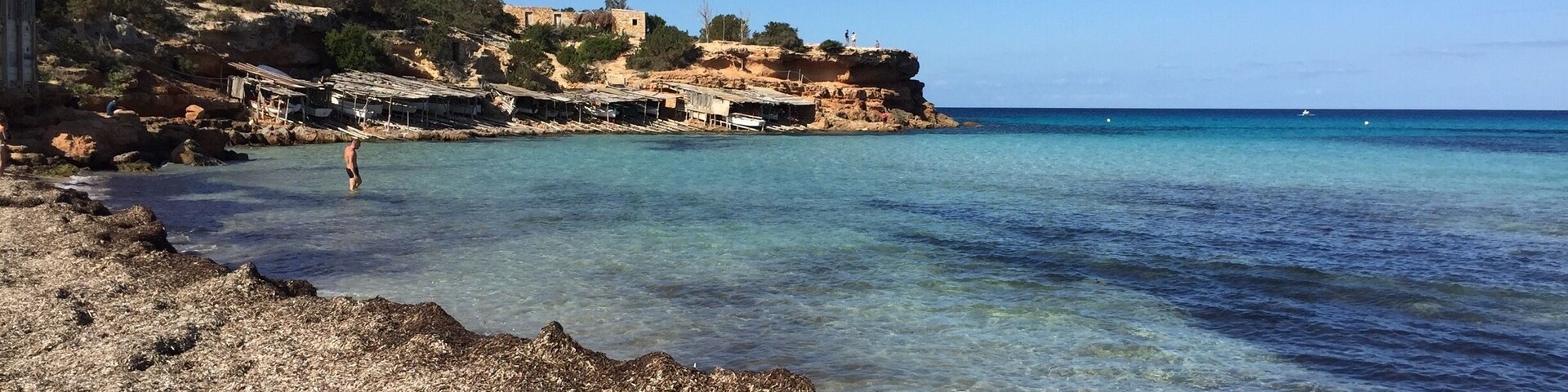 Una playa rocosa con agua turquesa cristalina, un acantilado con edificios y un velero a lo lejos.