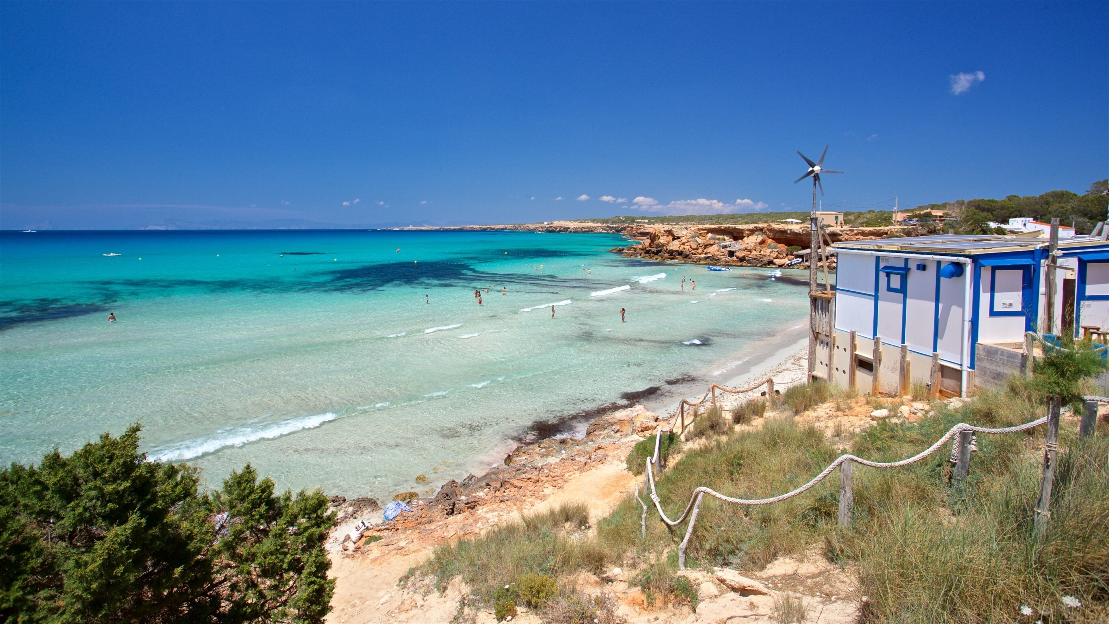 A beach with clear turquoise waters, people swimming and sunbathing, and a wind turbine in the background.