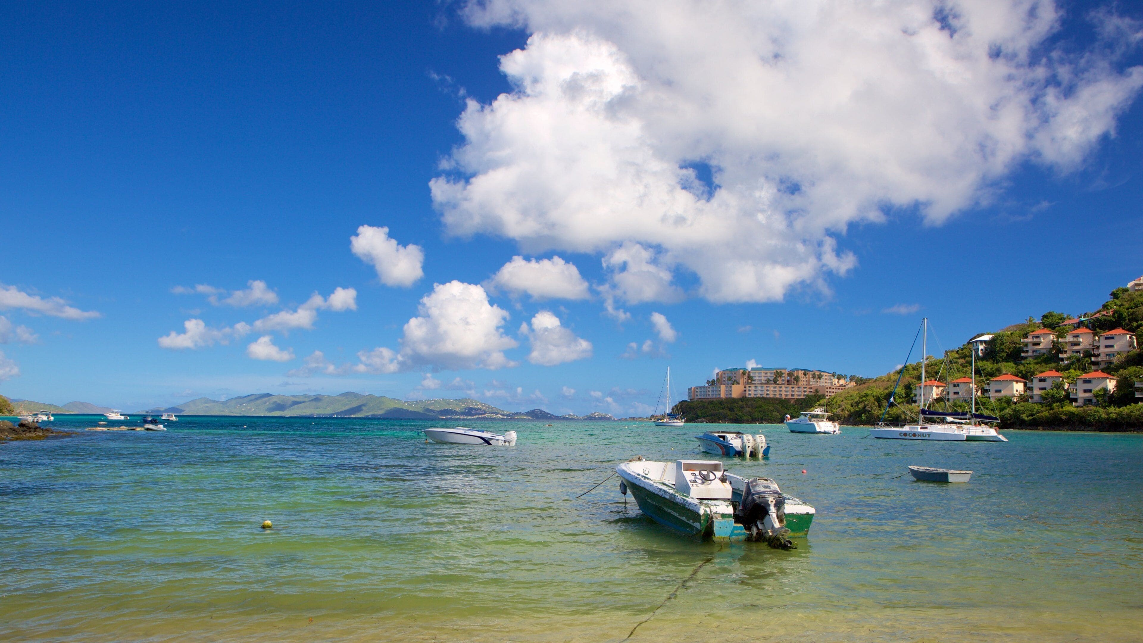 A coastal scene with boats anchored in clear turquoise waters, a distant mountain range, and a large resort building on a hillside.