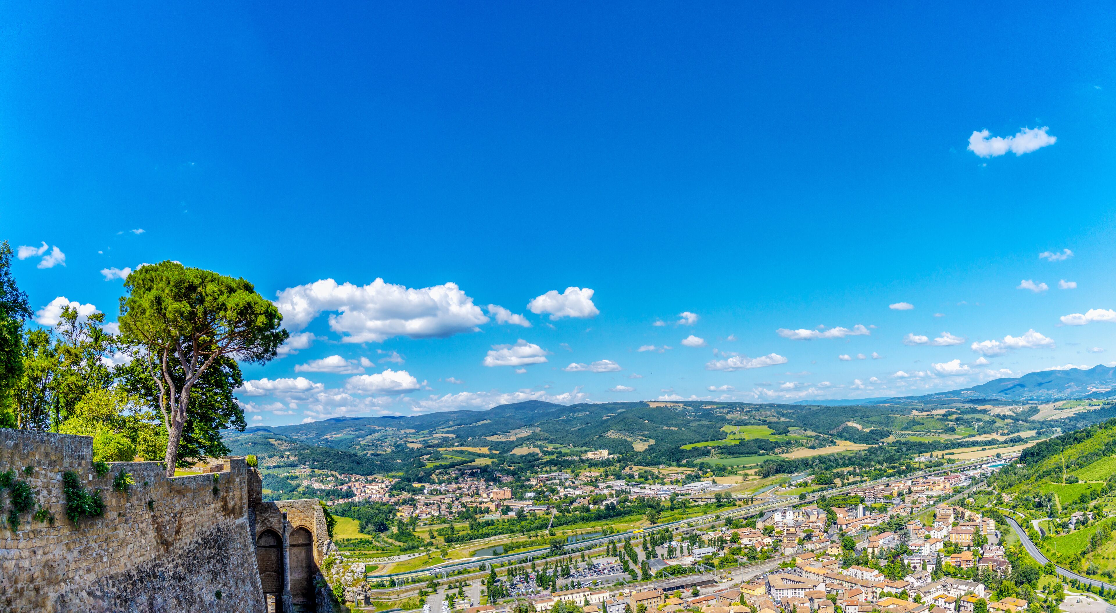 A panoramic view of a town with a prominent stone wall and a large tree on the left.