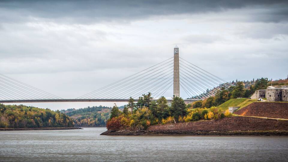 A suspension bridge over a river with autumn foliage and a historic building on the right.