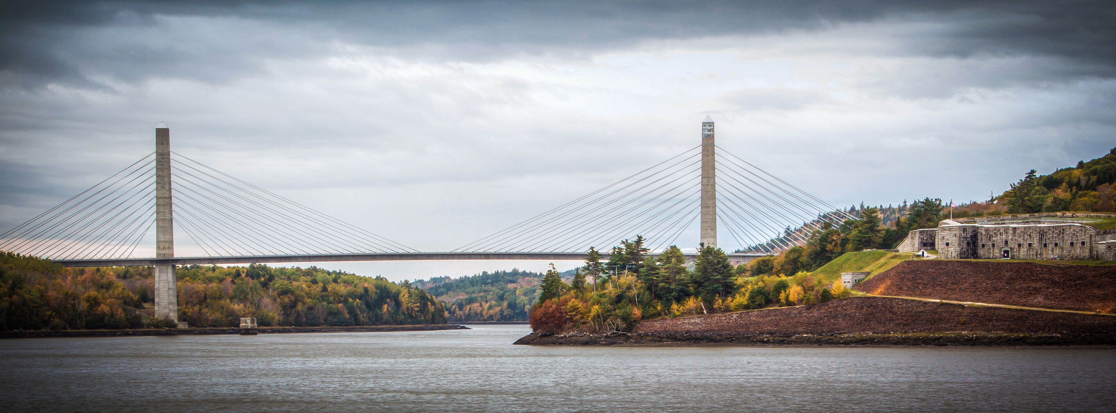 A suspension bridge over a river with autumn foliage and a historic building on the right.