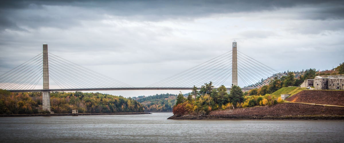 A suspension bridge over a river with autumn foliage and a historic building on the right.
