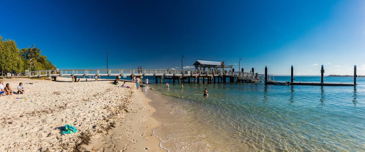 A beach with a wooden pier extending into the water, clear shallow waters, and a few people enjoying the area.