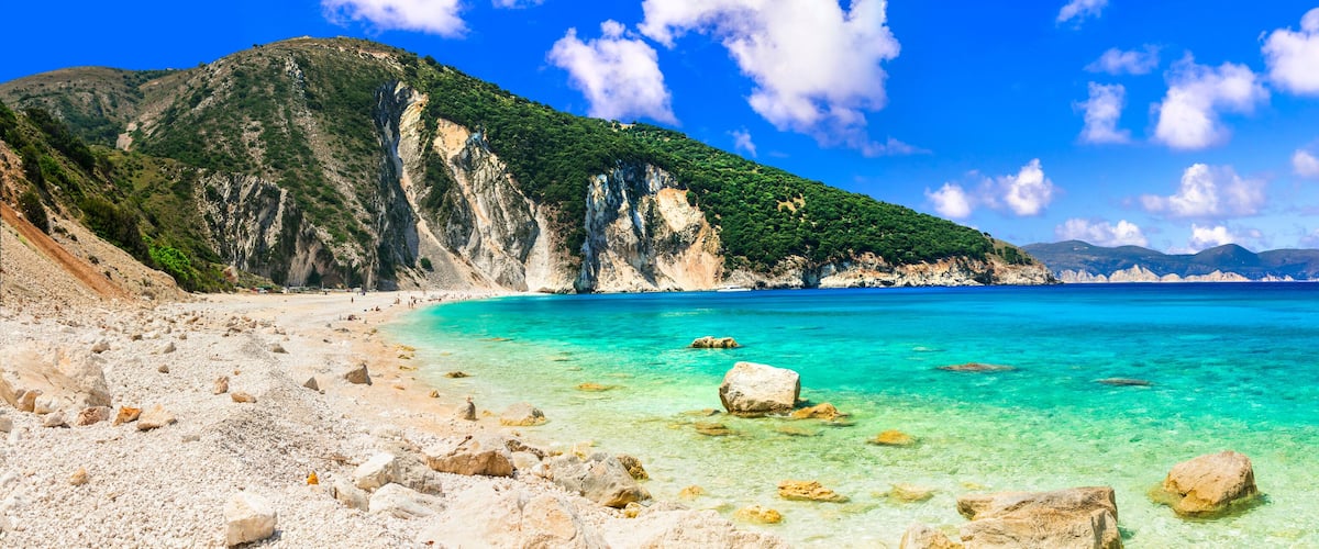 A coastal landscape with clear turquoise waters, rocky beach, and a mountainous backdrop.