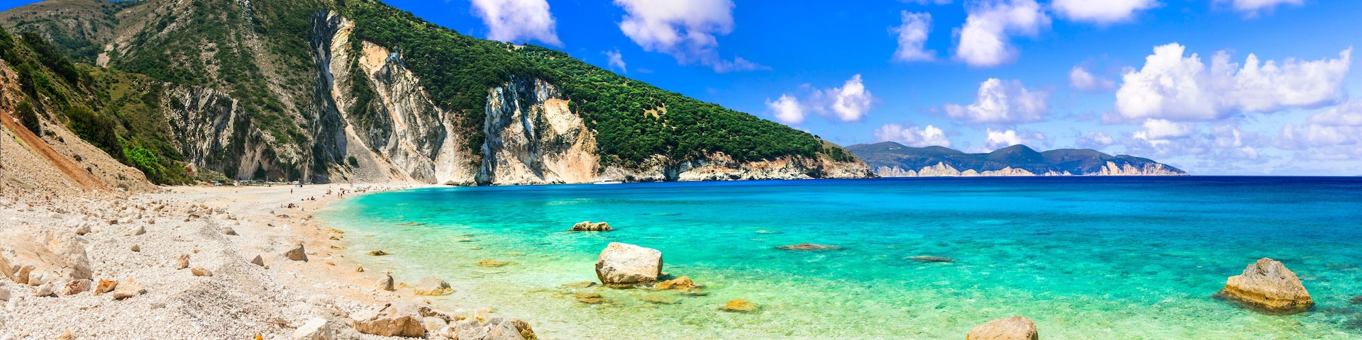 A coastal landscape with clear turquoise waters, rocky beach, and a mountainous backdrop.