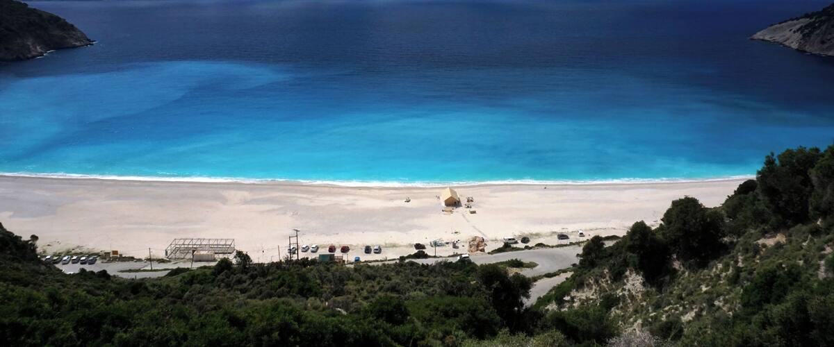 A coastal landscape with a beach, clear blue waters, and a mountainous backdrop.