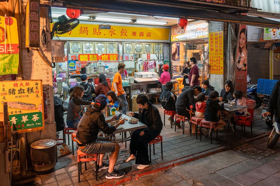Um mercado de comida de rua movimentado, com vários quiosques e pessoas a comer.