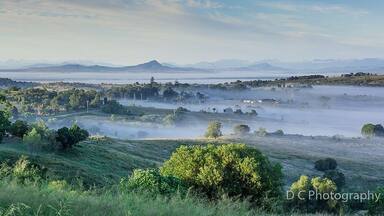 A misty landscape with rolling hills, scattered trees, and distant mountains.