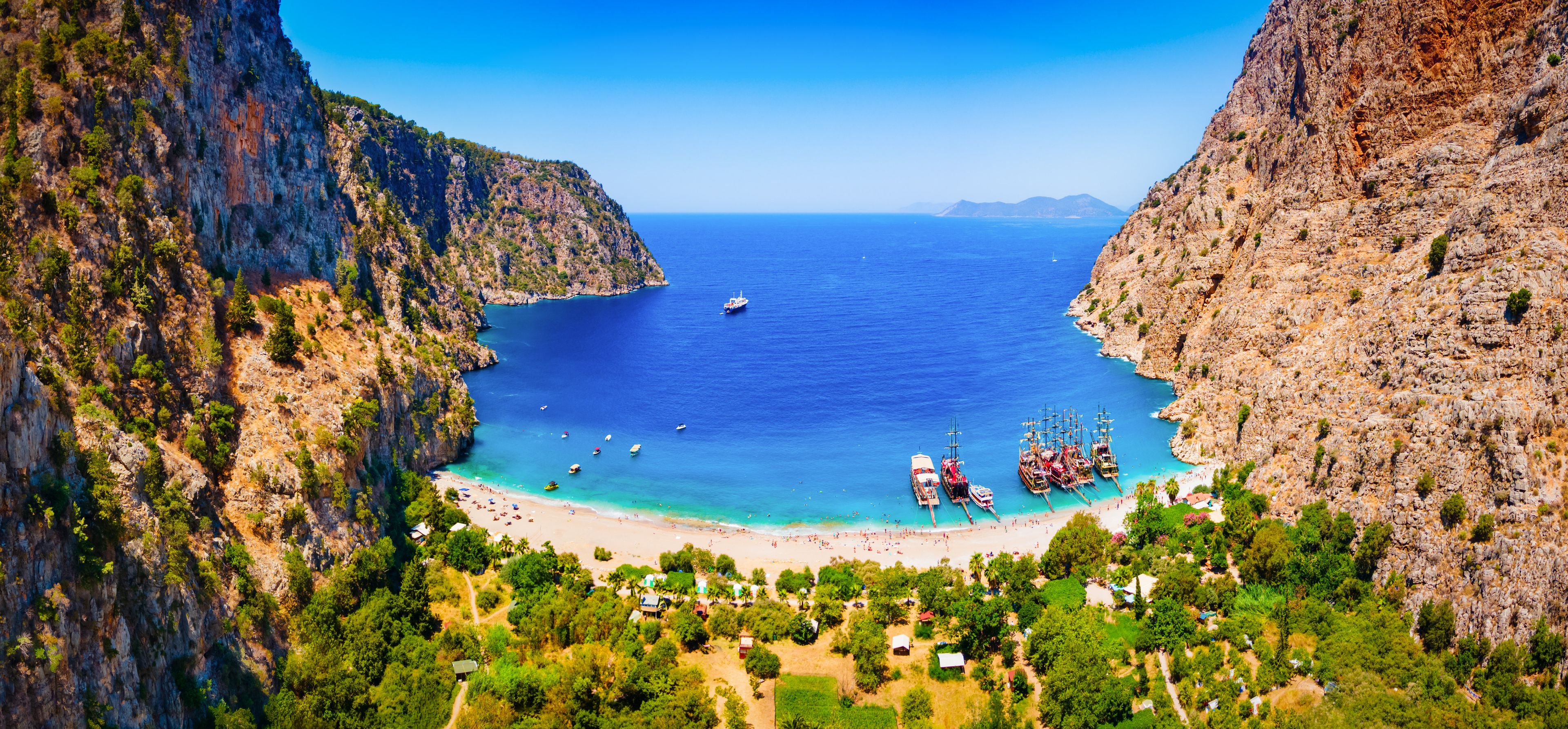 A coastal landscape with a beach, boats, and a mountainous backdrop.