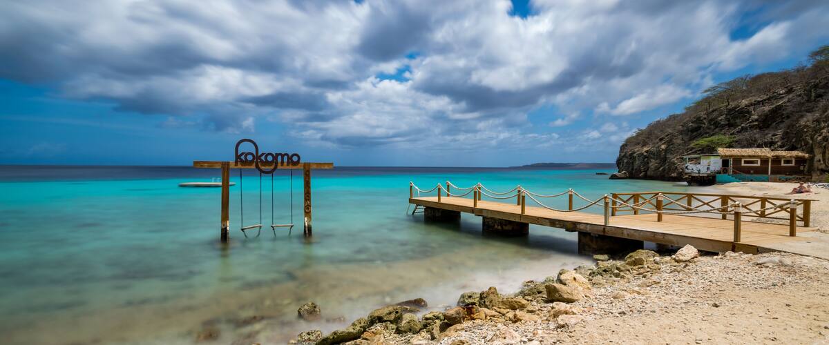 A wooden pier extending into turquoise waters, a sign reading 'kokomo,' and a rocky shoreline with a thatched-roof structure.