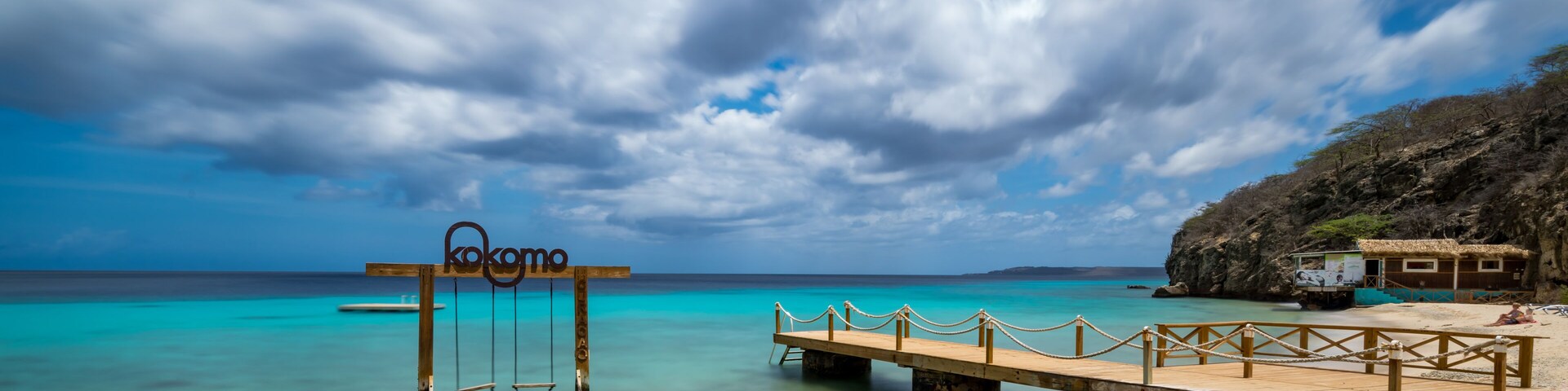 A wooden pier extending into turquoise waters, a sign reading 'kokomo,' and a rocky shoreline with a thatched-roof structure.