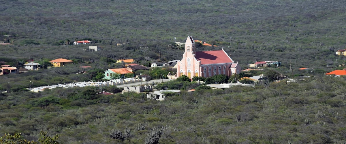 A church with a red roof and a bell tower, surrounded by houses and wind turbines on a hill.