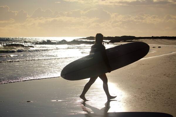 Eine Person trägt einen Surfbrett an einem Strand, im Hintergrund Wellen und ein Sonnenuntergang.