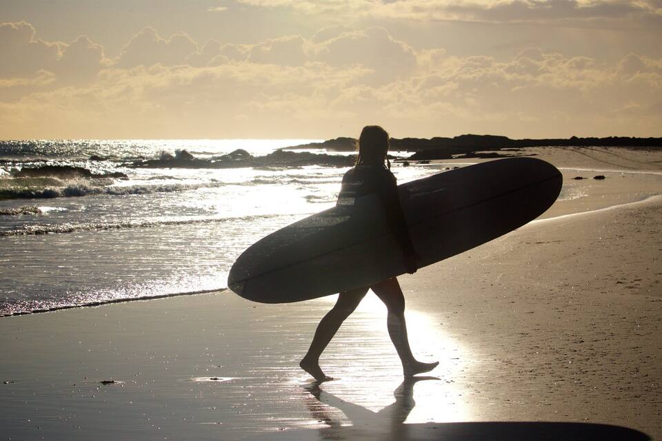A person carrying a surfboard on a beach with waves and a sunset in the background.
