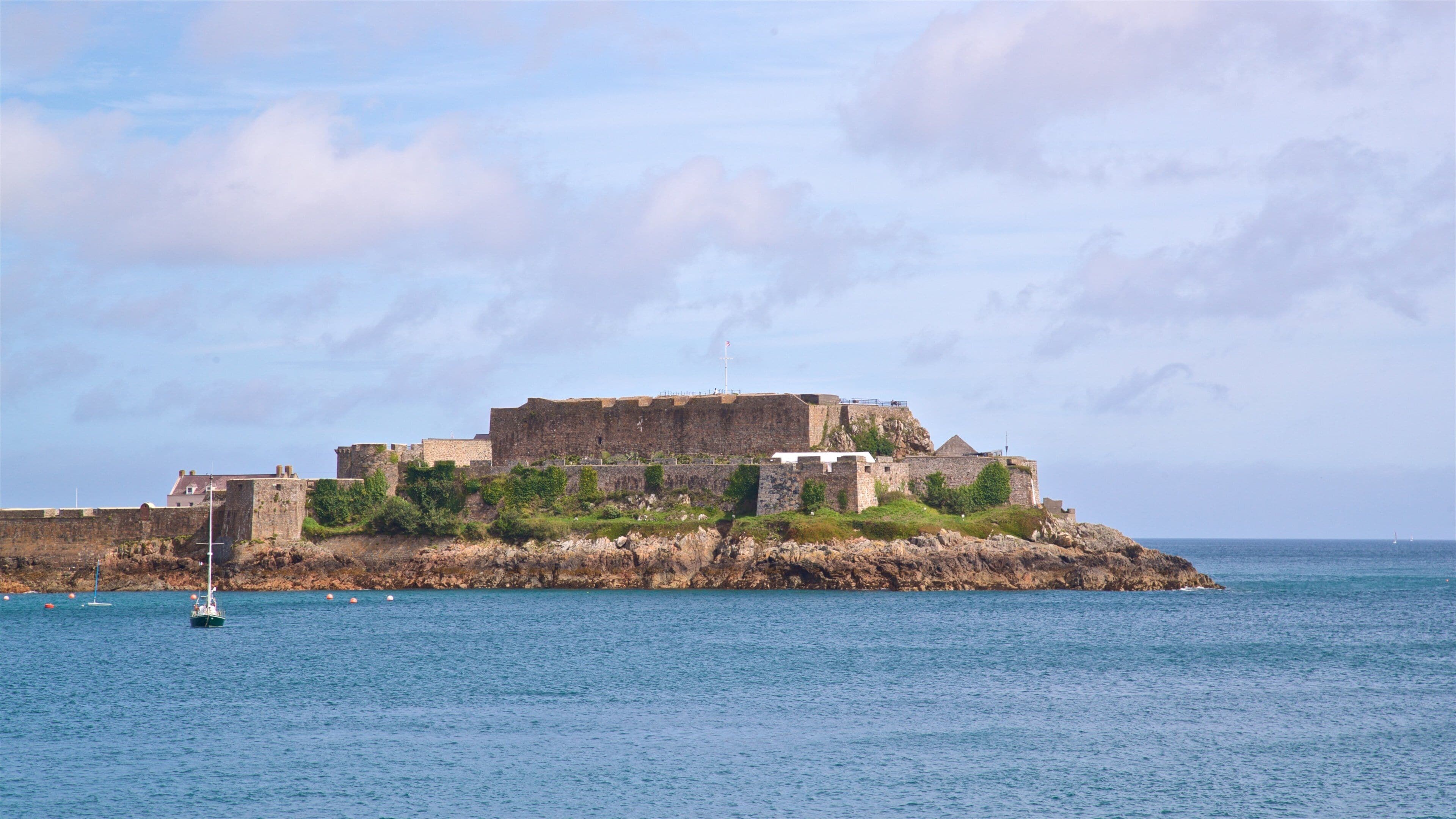 A coastal fortress with a flagpole, surrounded by water and a sailboat.