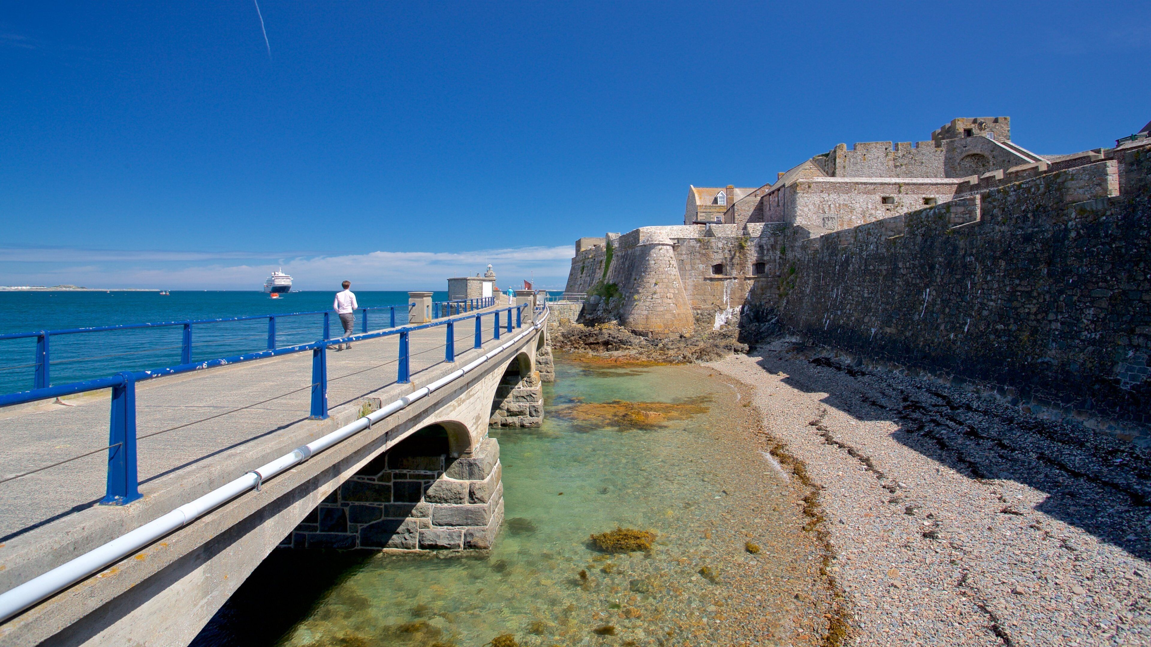 A stone bridge over clear water, a historic stone wall, and a person walking on the bridge.