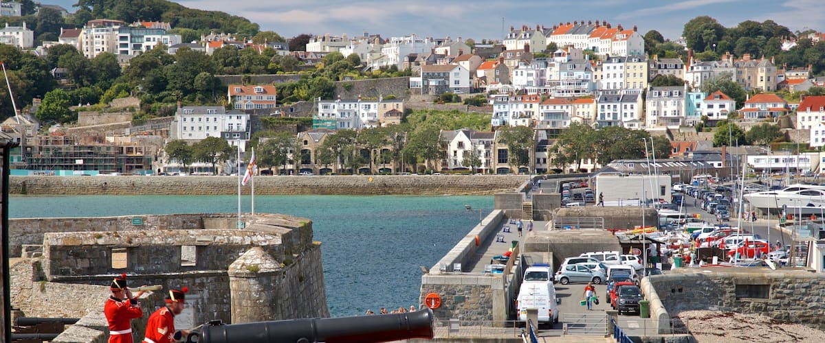 A coastal town with a historic cannon, stone walls, and a marina.