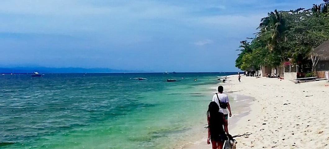 Una playa arenosa con agua turquesa cristalina, algunas embarcaciones a lo lejos y una persona caminando hacia el agua.