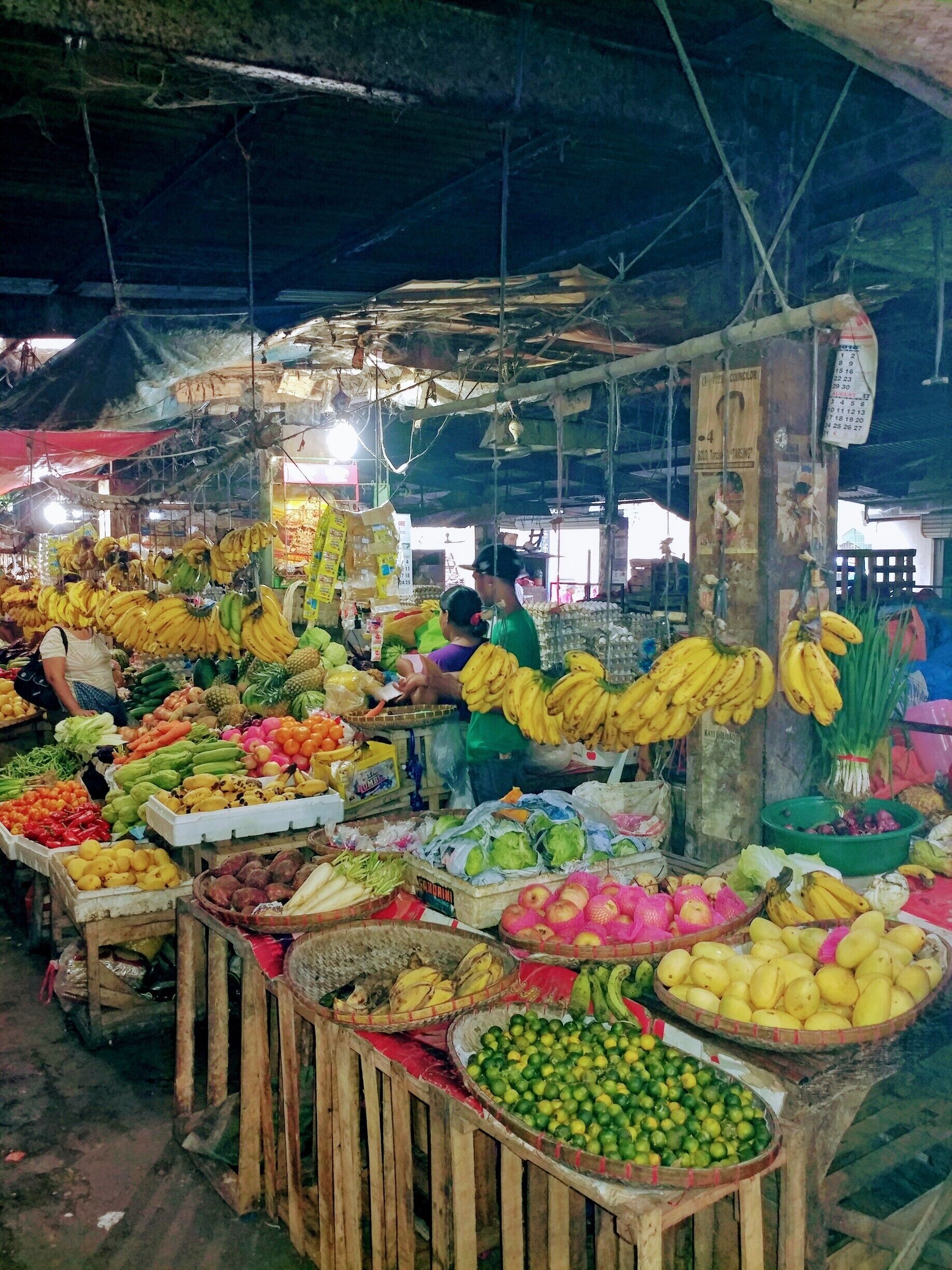 Um mercado interno movimentado, com diversas frutas e legumes expostos em caixas e cestos de madeira.