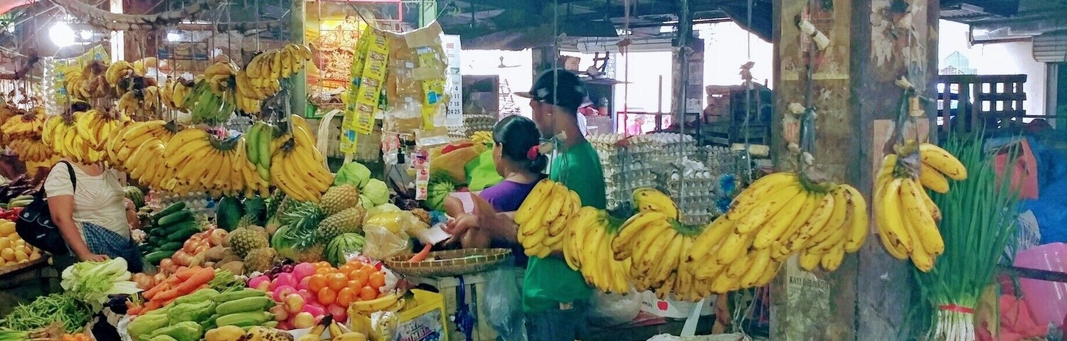 Um mercado interno movimentado, com diversas frutas e legumes expostos em caixas e cestos de madeira.