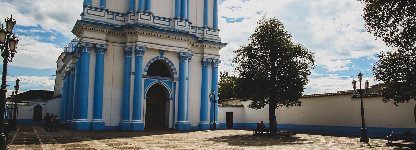 Una iglesia histórica con arquitectura azul y blanca, un patio empedrado y un árbol grande.